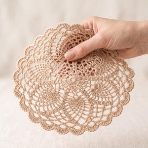 May include: A round, beige crochet doily featuring an intricate, openwork pineapple pattern with a scalloped edge. The circular textile is crafted with fine thread, displaying a detailed, symmetrical design radiating from the center outward.
