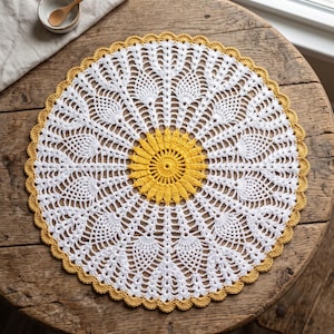 May include: A round, handmade crochet doily featuring a vibrant yellow center with a sunburst pattern, surrounded by intricate white pineapple-style lace motifs, and finished with a scalloped yellow border edge.