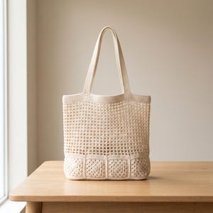 Puede incluir: Un bolso tote de ganchillo color crema con un patrón de malla abierta en la sección superior y un diseño de motivos cuadrados sólidos en la base. El bolso incluye dos asas integradas y una estructura rectangular con base plana.