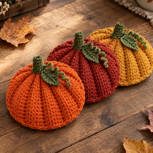May include: Three crocheted pumpkins in orange, dark red, and mustard yellow. Each pumpkin features a ribbed texture, a dark green stem, a single green leaf, and a small green curly vine attached to the top center of the rounded body.