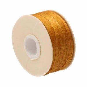 May include: A spool of golden-yellow cord wrapped around a white circular cardboard core. The cord has a textured, fibrous appearance and is wound tightly in multiple layers around the center of the spool, creating a cylindrical shape.