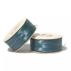 May include: Two spools of dark teal thread wound around circular cardboard cores. The thread has a smooth, slightly glossy finish and is wrapped tightly in a crisscross pattern around the center of each spool, leaving the edges exposed.