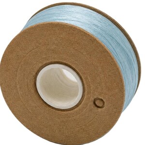 May include: A spool of light blue thread wound around a circular brown cardboard core. The spool features a white plastic center ring and a small circular hole punched into the side of the cardboard disc, showing the texture of the wound thread.