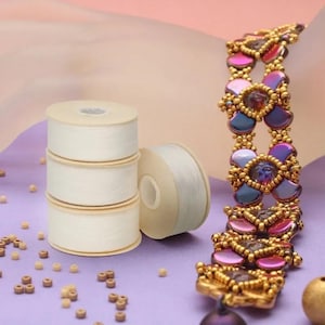 May include: A beaded bracelet featuring iridescent purple and blue petal-shaped beads framed by small gold metallic beads, positioned next to three stacked white spools of thread and scattered loose brown beads on a light purple surface.