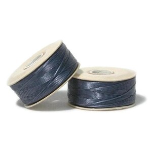May include: Two spools of dark blue waxed thread, each featuring a cylindrical shape with light-colored cardboard ends. The thread is tightly wound in a crisscross pattern around the center of the spools, creating a textured appearance.