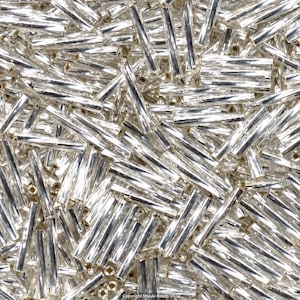 May include: A collection of numerous silver-colored glass bugle beads arranged in a dense, overlapping pattern. Each bead is long, thin, and cylindrical with a reflective, metallic finish. The text Copyright Miyuki Beads US is visible.