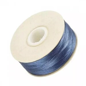 May include: A spool of blue cord featuring a smooth, braided texture wrapped around a circular, off-white cardboard core. The cord has a slight sheen and is wound tightly in multiple layers, creating a consistent, uniform appearance throughout the spool.