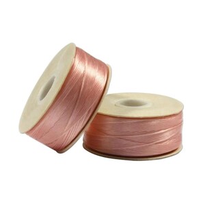 May include: Two spools of light pink, shiny, flat cord wrapped around circular white plastic cores. The cord has a smooth, satin-like texture and is wound tightly in a crisscross pattern around each spool, creating a uniform, reflective surface.