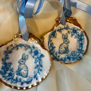 May include: Two seashell ornaments featuring blue rabbit and floral designs. The edges are trimmed in gold, and each has a light blue ribbon for hanging. The background is a soft, off-white fabric.