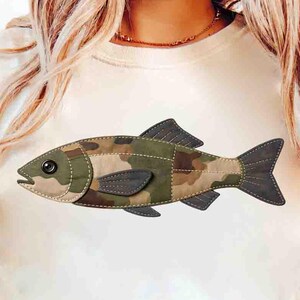 May include: Cream-colored t-shirt featuring a fish design. The fish is made of camouflage fabric with dark green and brown hues, and has a dark grey tail and fins. The fish is stitched onto the shirt with a light green thread.
