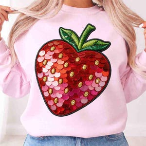 May include: A light pink sweatshirt featuring a large, sequined strawberry design. The strawberry is primarily red with pink and gold accents, and green embroidered leaves. The design is centered on the front of the sweatshirt.
