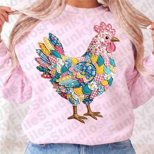 May include: A pale pink sweatshirt with a vibrant patchwork chicken appliqué. The chicken is adorned with a red comb and beak, and its body is crafted from diverse patterned fabrics in blue, yellow, and pink. A casual style.