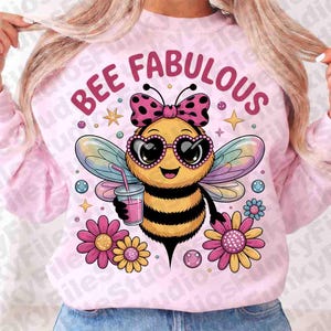 May include: Pale pink sweatshirt featuring a cartoon bee design. The bee has heart-shaped sunglasses and a bow, holding a drink. The text "BEE FABULOUS" is above the bee, with flowers and stars around it.