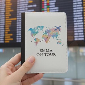 Könnte beinhalten: Ein weißer Reisepasshalter mit einem Weltkarten-Design in Aquarelltönen von Blau, Grün und Rosa. Eine blaue Flugzeuggrafik und der Text "EMMA ON TOUR" sind ebenfalls abgebildet. Der Halter wird vor einer Anzeigetafel gehalten.