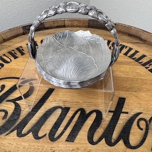 May include: A silver-colored metal basket with a handle adorned with floral details. The bowl of the basket has a leaf-like design. The basket is sitting on a clear acrylic stand, placed on a wooden surface.