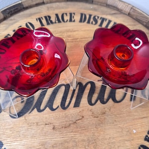 May include: Two red glass candle holders with a flower-like shape. Each holder has a central cup for a candle and sits on a clear acrylic stand. The holders are on a wooden surface.