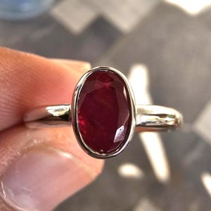 May include: A silver ring featuring a polished, oval-cut deep red gemstone set in a smooth bezel setting. The band is plain and metallic, showcasing a reflective surface. The gemstone displays a faceted texture that catches the light, centered within the metal frame of the ring.