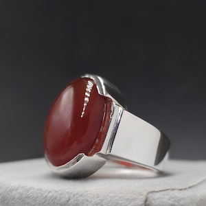 May include: A silver ring featuring a large, oval, polished red gemstone. The gemstone is set in a silver bezel and has a smooth, glossy surface. The ring band is wide and rectangular, with a polished finish. The ring is displayed on a white surface.