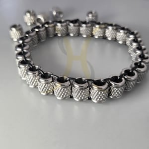 Puede incluir: Una pulsera plateada con cuentas cilíndricas texturizadas ensartadas en un cordón negro. Varias cuentas sueltas están esparcidas alrededor de la pulsera. La pulsera tiene un diseño moderno e industrial.