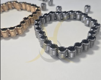 May include: Two bracelets, one gold-toned and one silver-toned, each composed of cylindrical beads with a textured, ridged design. Several loose beads are scattered nearby. The bracelets are displayed on a white surface.