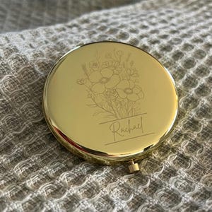 May include: A round, gold-colored compact mirror with a floral design and the name "Rachael" engraved on the front. The compact has a small clasp and is set against a textured background.