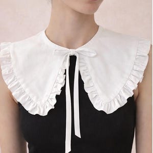Op de afbeelding: Een witte, gefranjerde kraag met een strik, gedragen over een zwarte top. De kraag heeft een afgeronde vorm en een delicaat ontwerp. De strik hangt naar voren en voegt een vleugje elegantie toe. Een modeaccessoire.