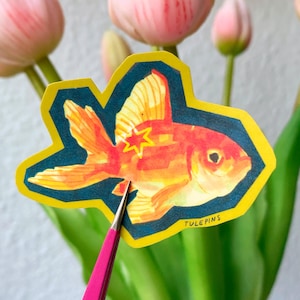 Puede incluir: Una pegatina de pez dorado con borde amarillo y fondo azul oscuro. El pez dorado es naranja y tiene una estrella en un lado. La pegatina está sujeta por unas pinzas rosas frente a tulipanes rosas y hojas verdes. El texto "TULEPINS" es visible.