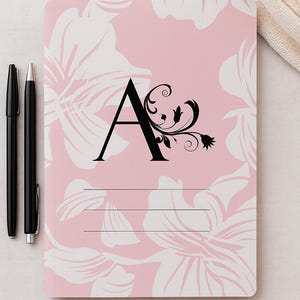 Op de afbeelding: Een roze notitieboek met een bloemenpatroon en de zwarte letter "A", versierd met een decoratief krulwerk. Twee zwarte pennen zijn links geplaatst. Het notitieboek heeft drie horizontale lijnen om op te schrijven.