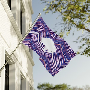 May include: A flag with a blue and white striped pattern and a white silhouette of a bear. The flag is mounted on a silver pole and is waving in the wind against a backdrop of a building and a tree.
