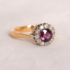 Peut inclure: Une bague en or avec une pierre précieuse centrale ronde d'un violet profond, entourée d'un halo de petites pierres claires taillées en rond. L'anneau est lisse et poli, passant d'une tige en or à un sertissage aux tons argentés.