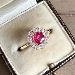 May include: A gold ring featuring a central round faceted red gemstone surrounded by a halo of ten clear round faceted stones. The ring is set in a yellow gold band and is displayed inside a cream-coloured velvet-lined jewellery box.