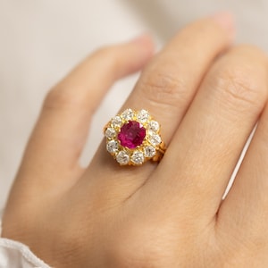 Puede incluir: Un anillo de oro con un rubí central de talla cojín en color rojo rosado intenso, rodeado por un halo de diez diamantes blancos de talla brillante redonda. El engaste es de oro amarillo y presenta un diseño clásico de racimo.