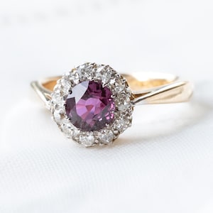 Peut inclure: Une bague en or avec une pierre centrale ronde d'un violet profond entourée d'un halo de petites pierres rondes transparentes. L'anneau est lisse et s'affine vers le sertissage, fabriqué en métal argenté pour maintenir les pierres.