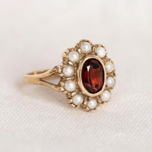 May include: A gold ring featuring a central oval-cut deep red garnet gemstone surrounded by a halo of small white pearls. The metal band has a split shank design, and the setting is crafted with a scalloped edge around the central stone.