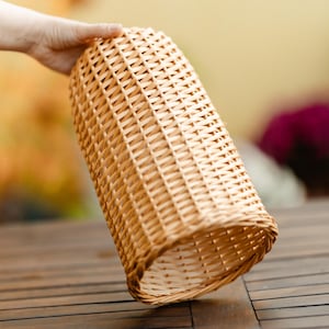 May include: A woven wicker basket with a rounded, cylindrical shape. The basket is held by a hand and is made of light brown wicker. The weave pattern is visible, and the basket appears to be empty. The background is blurred.