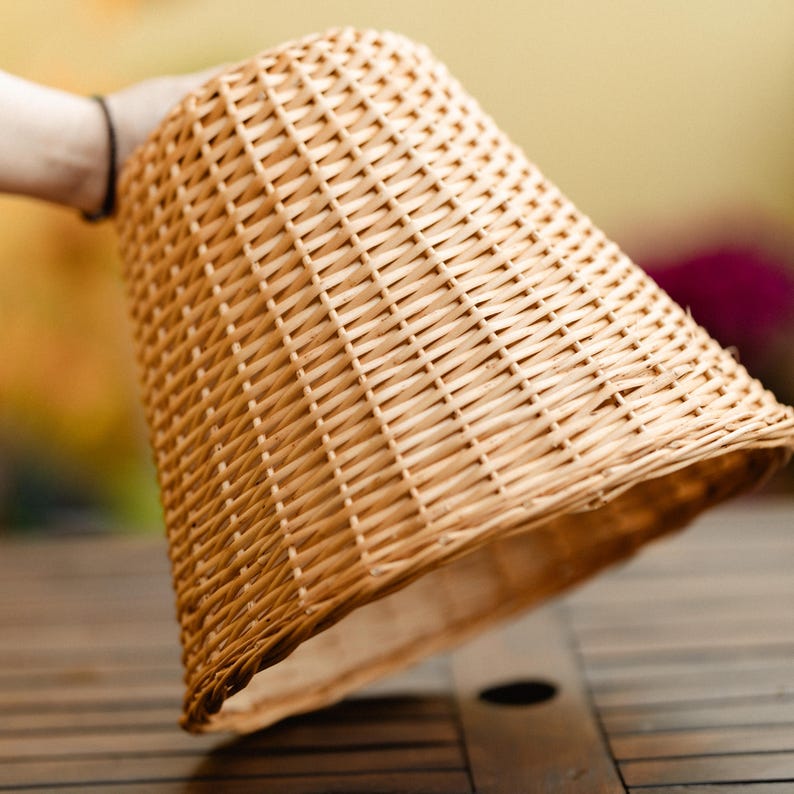 May include: A woven wicker lampshade in a conical shape. The lampshade is held by a person's hand and is made of light brown wicker. The weave pattern is visible, and the lampshade is slightly tilted.