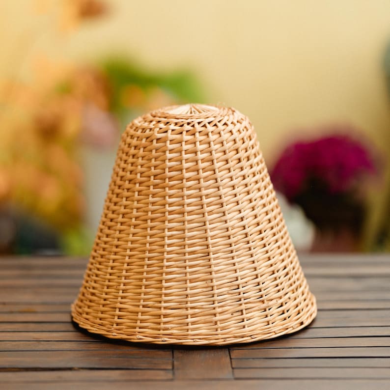 May include: A light brown, woven wicker lampshade with a conical shape. The lampshade is made of tightly woven, natural-colored wicker and sits on a dark wooden surface. The background is blurred, with hints of greenery and flowers.