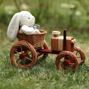 May include: A white plush bunny sits in a woven basket attached to a toy tractor. The tractor is made of wood and woven wicker, with red accents. The wheels are made of woven wicker. The toy is set on a bed of green grass.