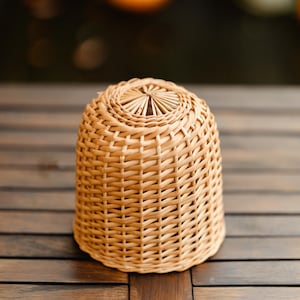 May include: A woven wicker basket with a rounded, bell-like shape. The basket is made of light brown wicker and has a tightly woven pattern. The top of the basket has a decorative, sunburst-like design.