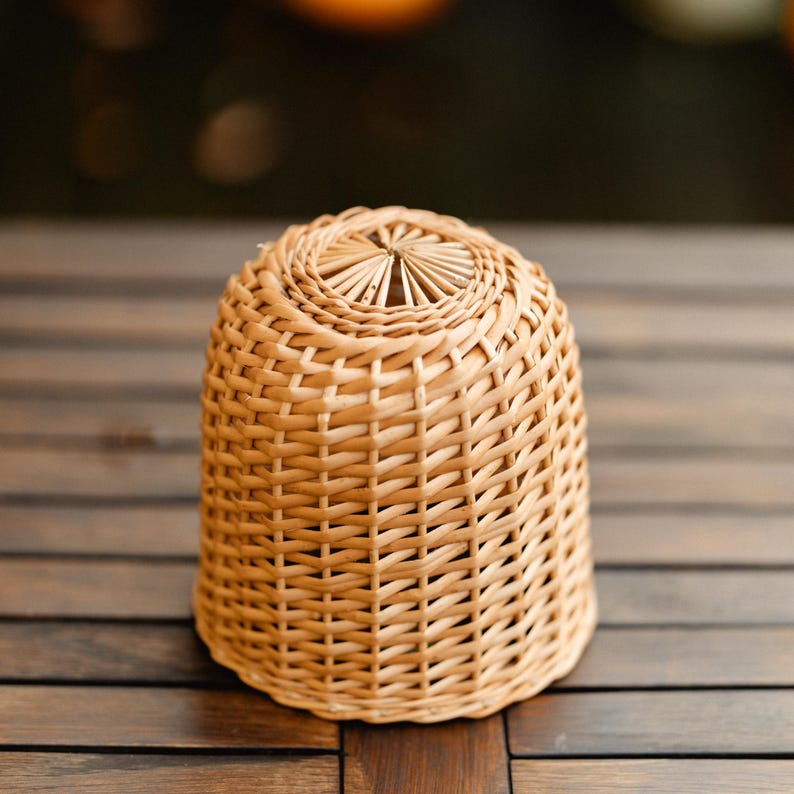 May include: A woven wicker basket with a rounded, bell-like shape. The basket is made of light brown wicker and has a tightly woven pattern. The top of the basket has a decorative, sunburst-like design.