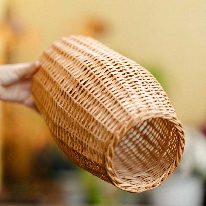 May include: A woven wicker basket with a cylindrical shape. The basket is made of light brown wicker and has a textured surface. The basket is held by a person's hand.