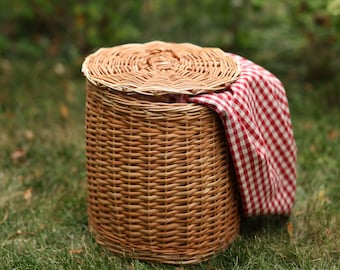 May include: A round, woven wicker basket with a lid, set on green grass. A red and white checkered cloth is draped over the side. The basket is a natural brown color, and the lid is slightly ajar.