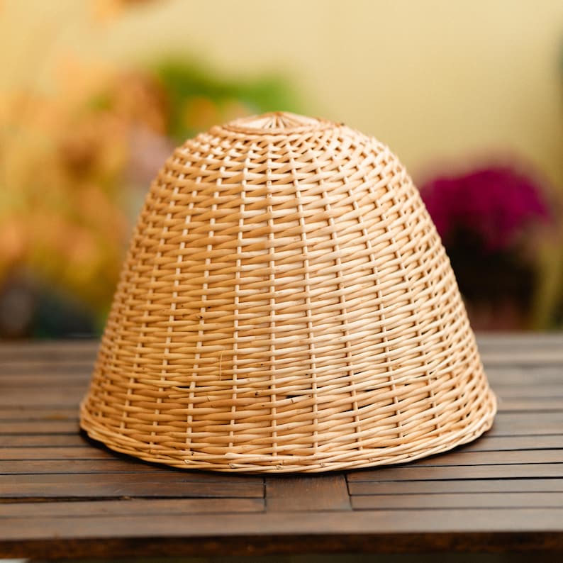 May include: A light brown, woven wicker lampshade with a rounded top and a flared, conical shape. The lampshade is made of tightly woven, natural-colored wicker and sits on a dark wooden surface. The background is blurred with hints of green and purple.