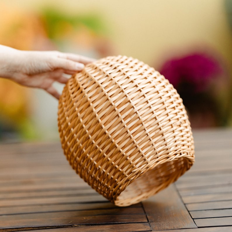 May include: A woven wicker basket with a rounded shape. The basket is made of light brown wicker and has a textured surface. The basket is held by a hand, with a blurred background.