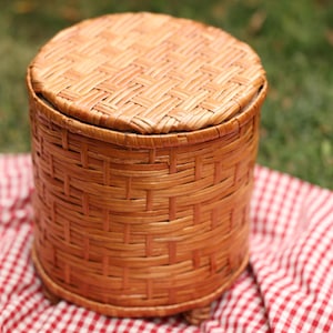 May include: A round, woven wicker basket with a lid. The basket is a warm brown color and has a textured surface. The lid fits snugly on top. The basket is sitting on a red and white checkered cloth.