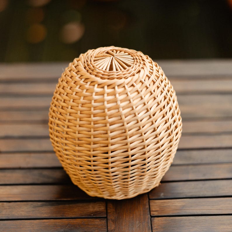 May include: A woven basket with an egg-like shape. The basket is made of light brown wicker and has a tightly woven pattern. The top of the basket has a circular design. The basket is sitting on a dark brown wooden surface.