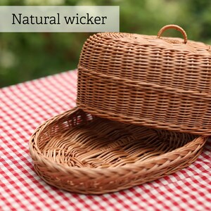 Op de afbeelding: Een natuurlijke rieten mand met deksel, geplaatst op een rood-wit geruite tafelkleed. Het deksel heeft een handvat. De tekst "Natural wicker" is zichtbaar.