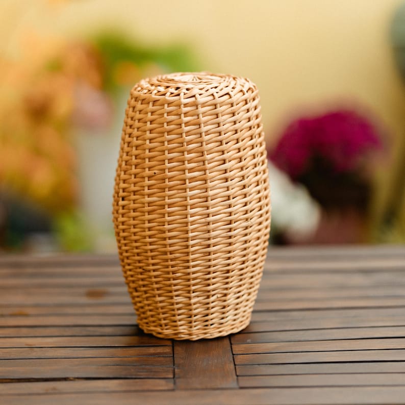 May include: A woven wicker vase in a light brown color. The vase has a rounded, oval shape with a woven pattern. The vase is sitting on a dark brown wooden table.