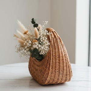 May include: A woven wicker basket with a handle, filled with dried flowers and greenery. The basket is a light brown color and has a curved shape. The flowers are a mix of white, beige, and green, creating a natural and rustic aesthetic.