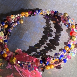 May include: A multi-strand bracelet with a wrap-around design. The bracelet features a variety of colorful beads, including clear, gold, purple, blue, and pink. The beads are arranged in a spiral pattern, creating a textured and eye-catching accessory.